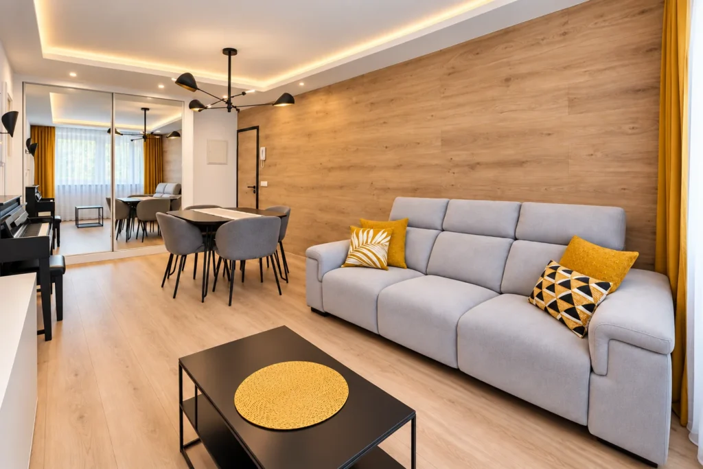 Salón moderno con sofá gris, cojines amarillos y pared revestida de madera, abierto a la zona de comedor.