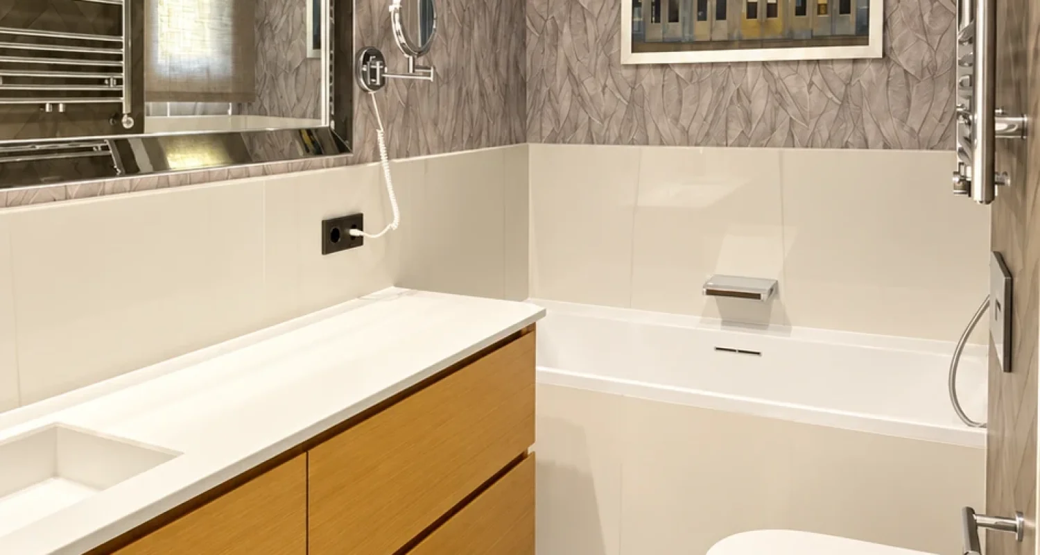 Baño principal reformado en ático de Aviador Zorita, Madrid, con mueble de madera suspendido, espejo iluminado y revestimiento gris.