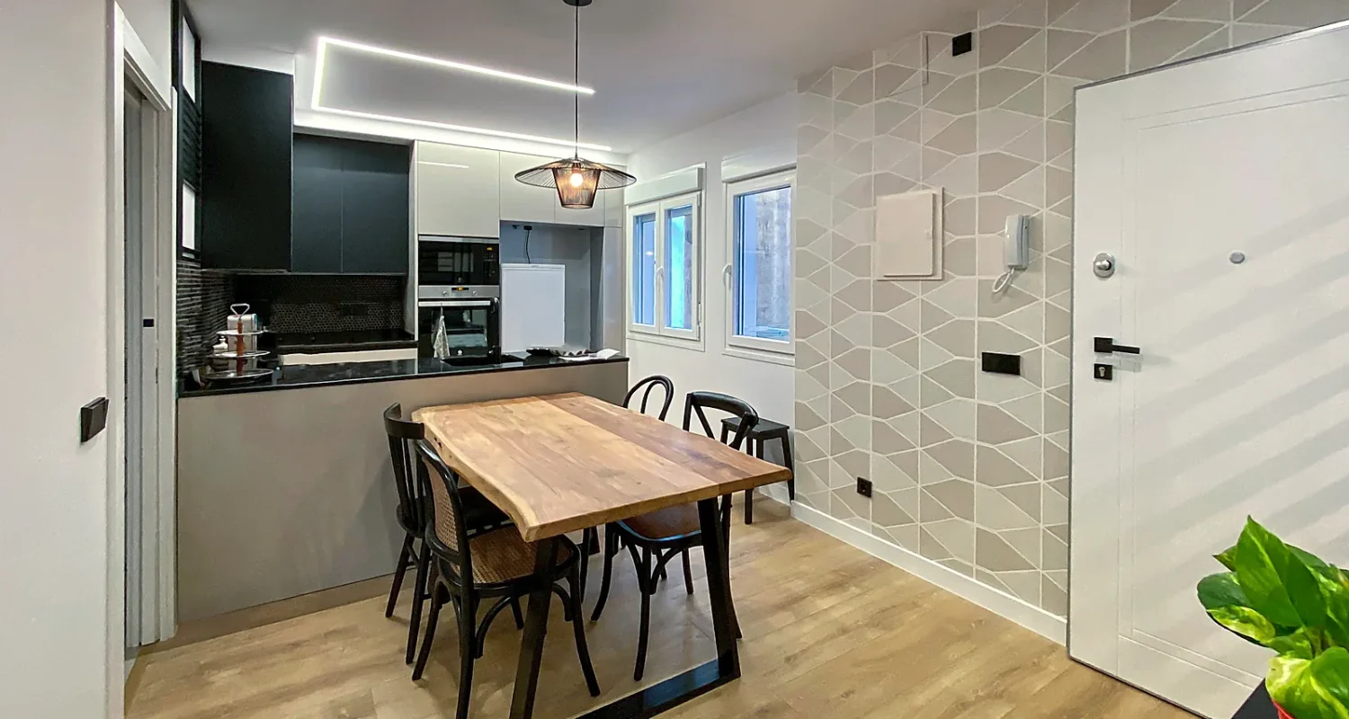 Cocina abierta al comedor en reforma integral de piso en calle Tarragona, Madrid, con mesa de madera, mobiliario moderno y zona de entrada.