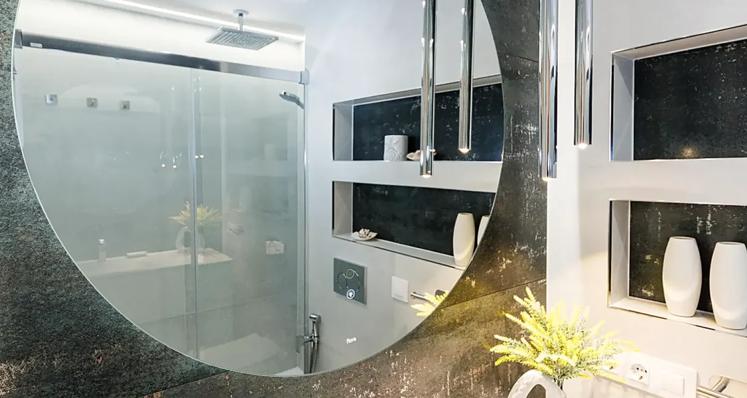 Baño reformado en Mejorada del Campo, Madrid, con espejo redondo, lavabo blanco y revestimiento oscuro efecto mármol.